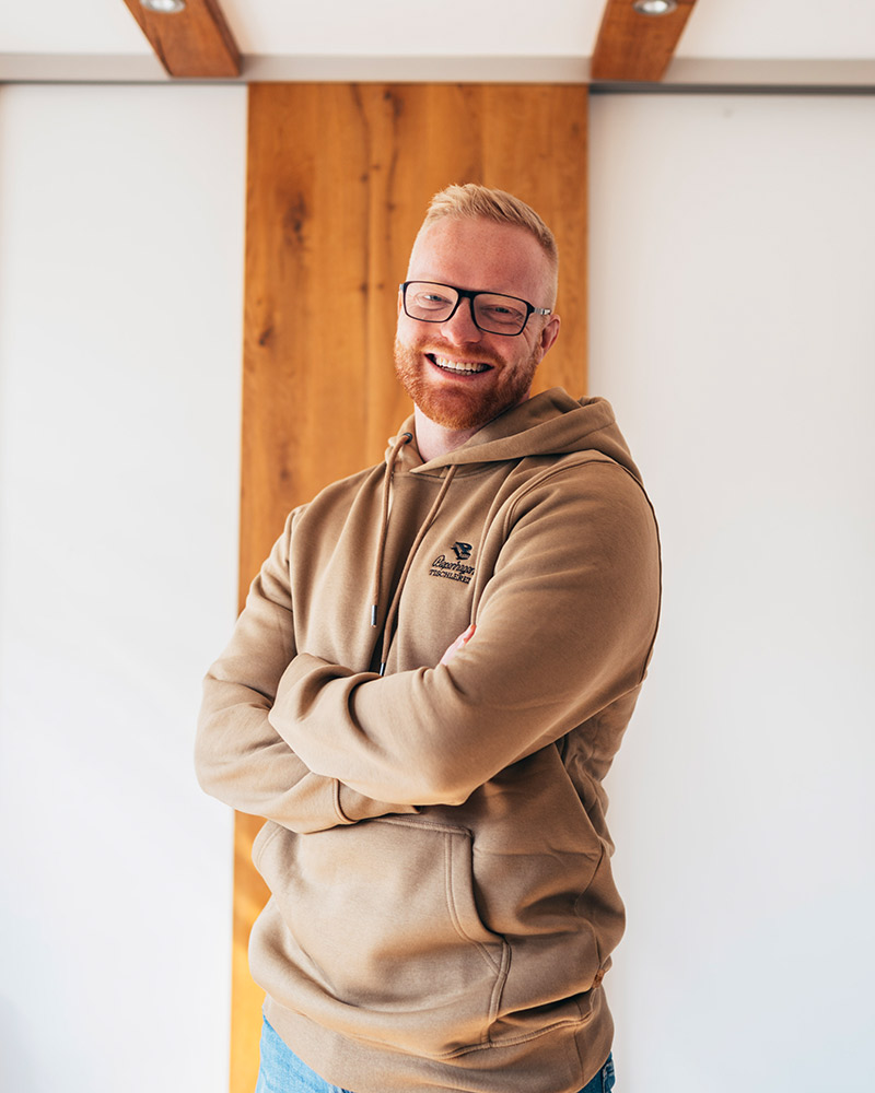 Ein lächelnder Mann mit hellem Bart trägt eine beige Kapuzenjacke und Brille