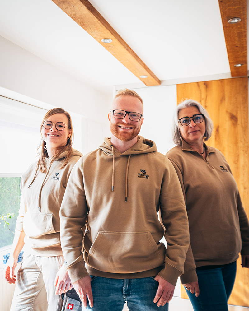 Drei lächelnde Personen mit Brillen tragen beige Hoodies in einem hellen Raum