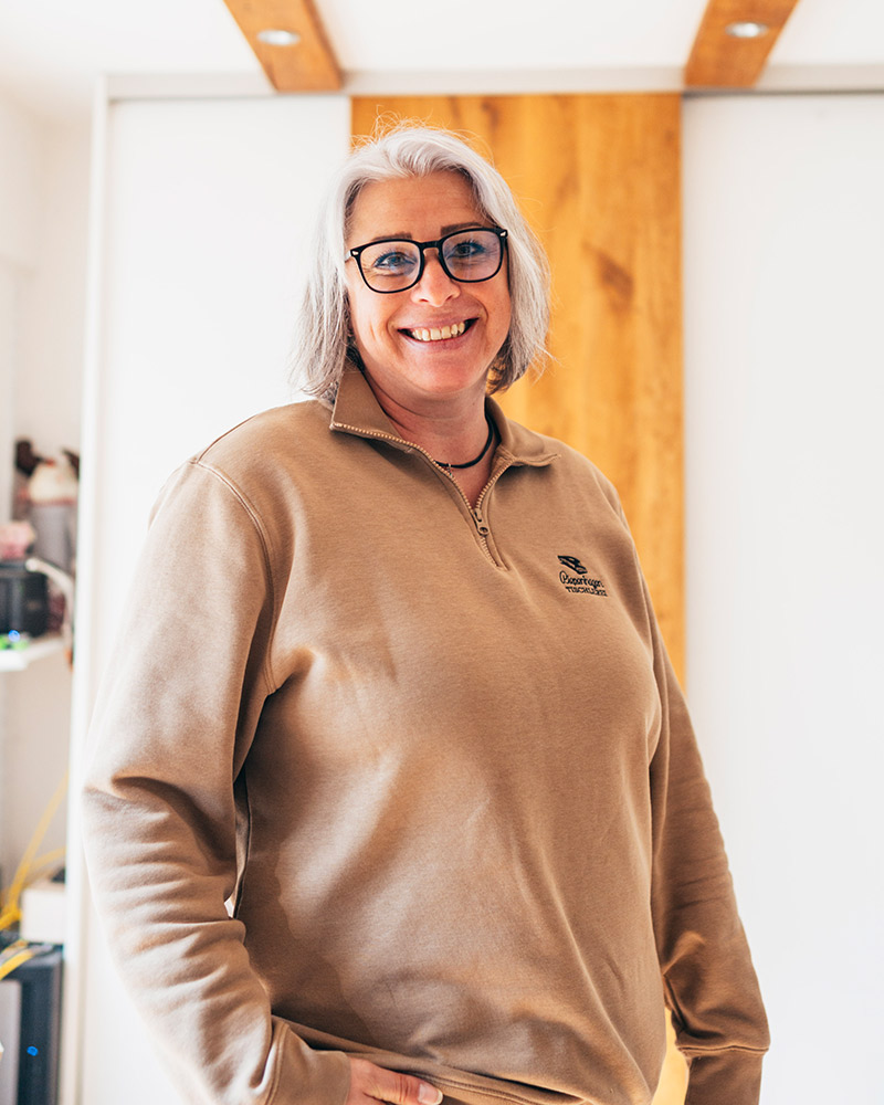 Eine grauhaarige Frau mit Brille lacht freundlich und trägt einen beigen Pullover