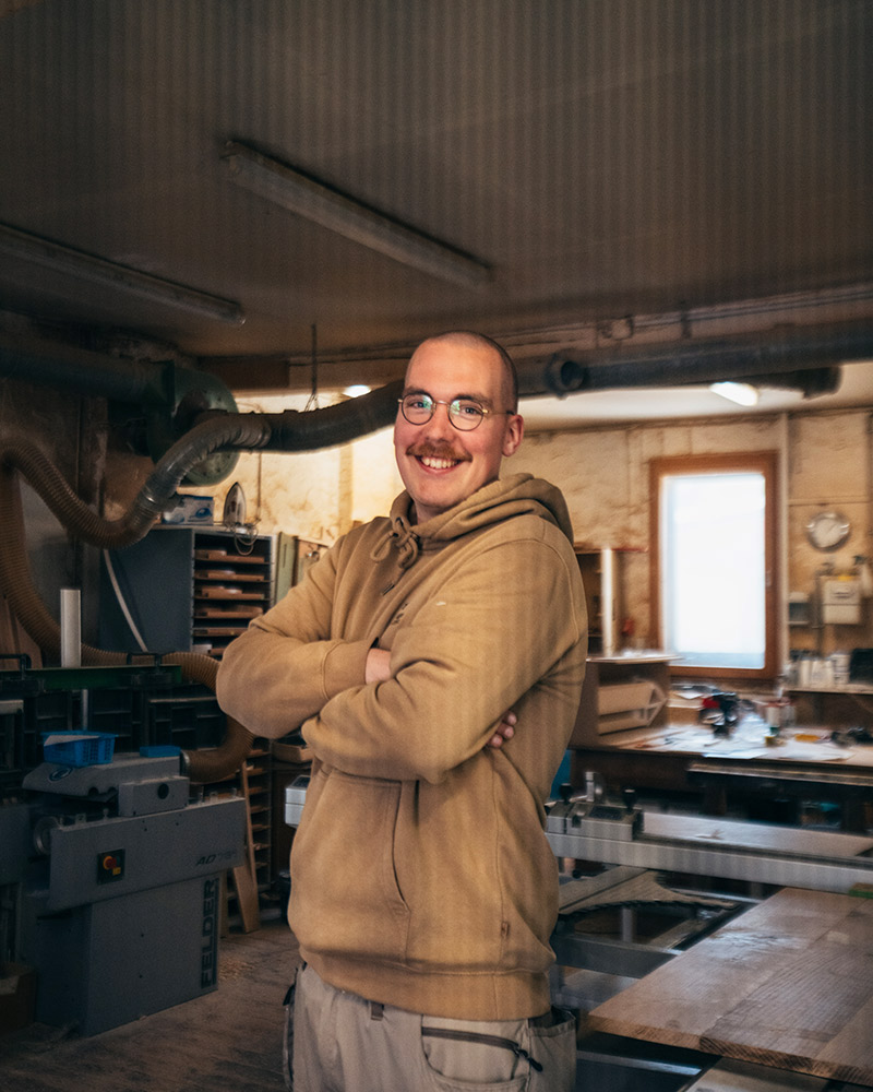 Ein lächelnder Mann mit Brille und Schnurrbart steht in einer hellen Werkstatt